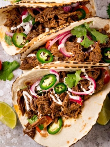 Square image of beef barbacoa on soft tacos topped with red onion and jalapeno slices