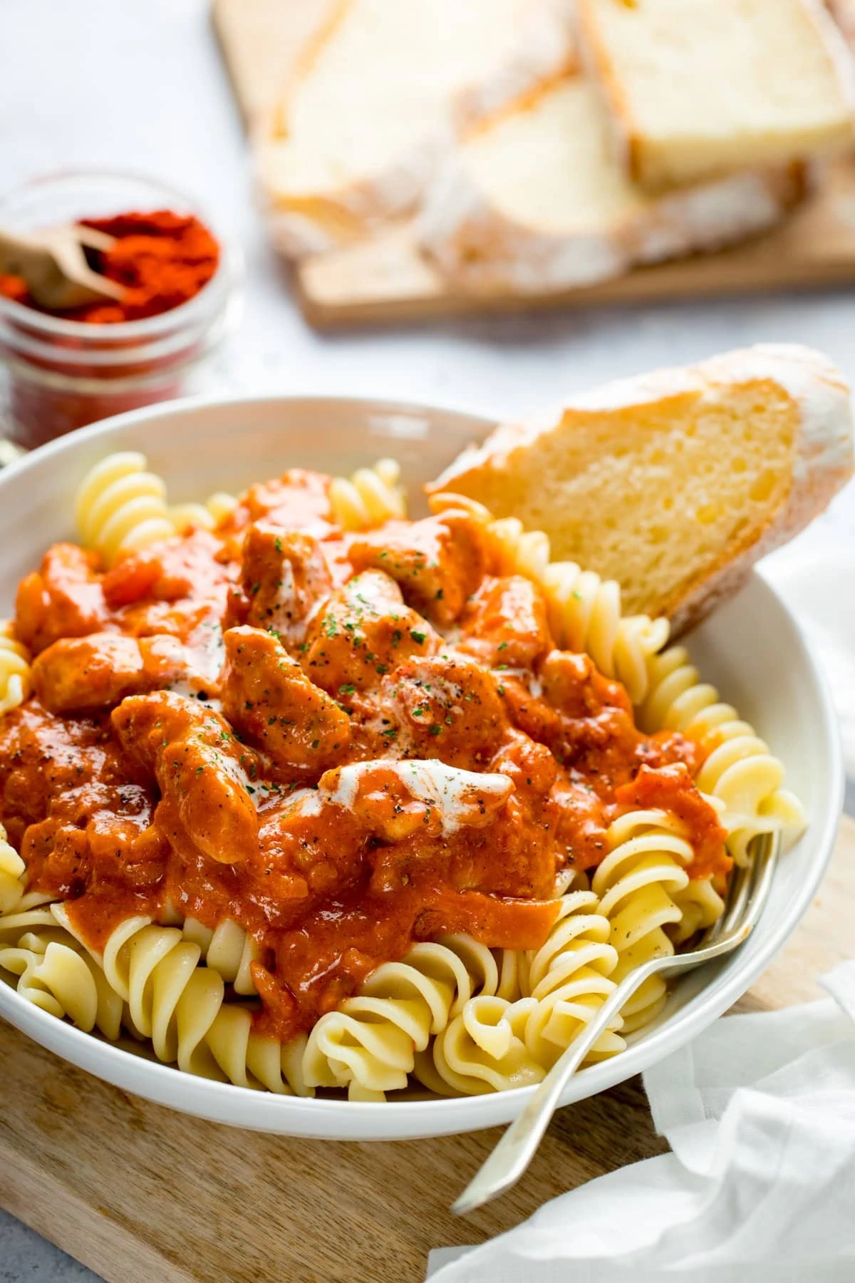 White bowl on a light background filled with pasta and chicken paprikash. Slices of bread and a jar of paprika at the top of the image.