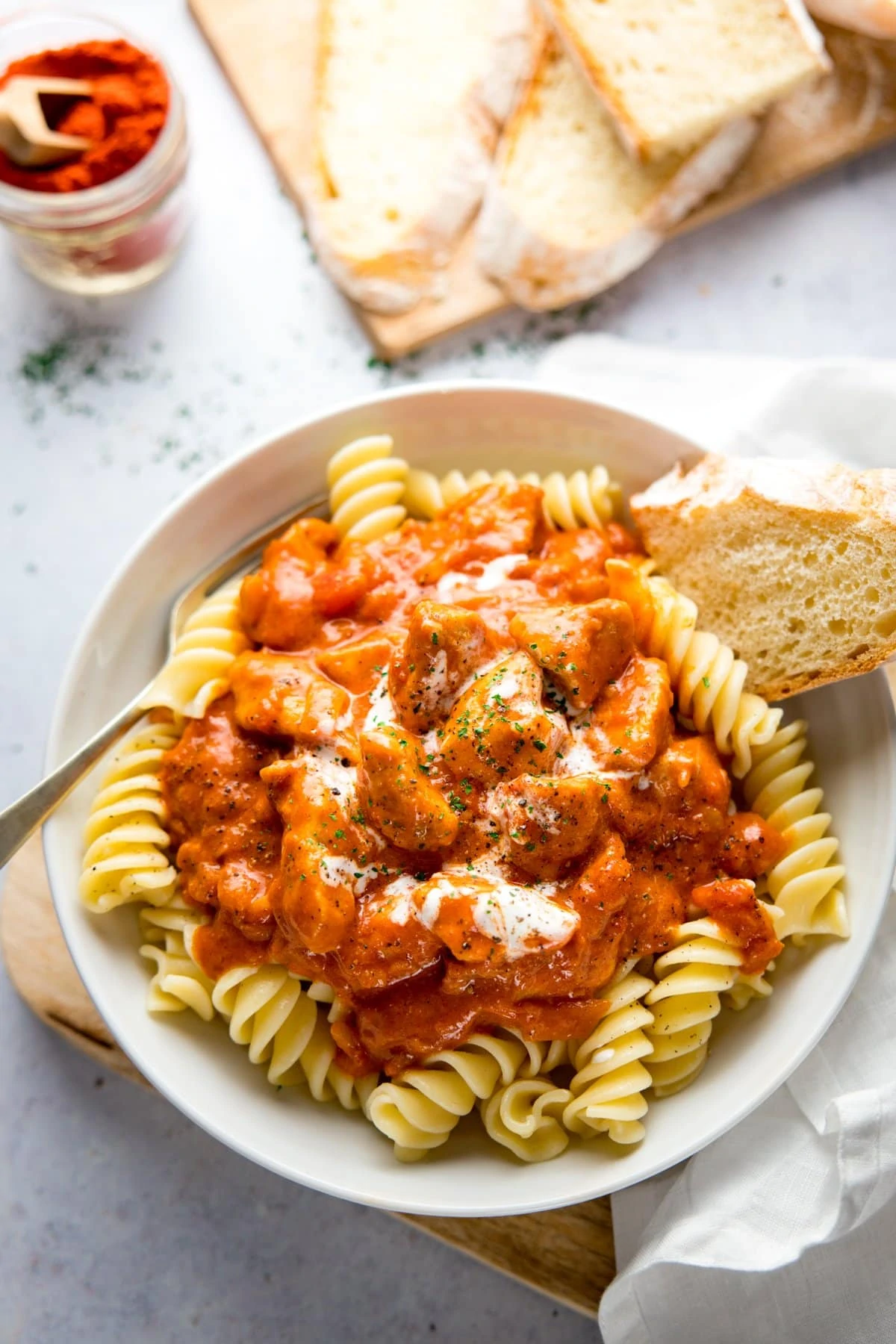 Overhead image of a white bowl filled with chicken paprikash, pasta and a piece of bread. Bowl is on a light background and there is more bread a napkin and jar of paprika in shot.