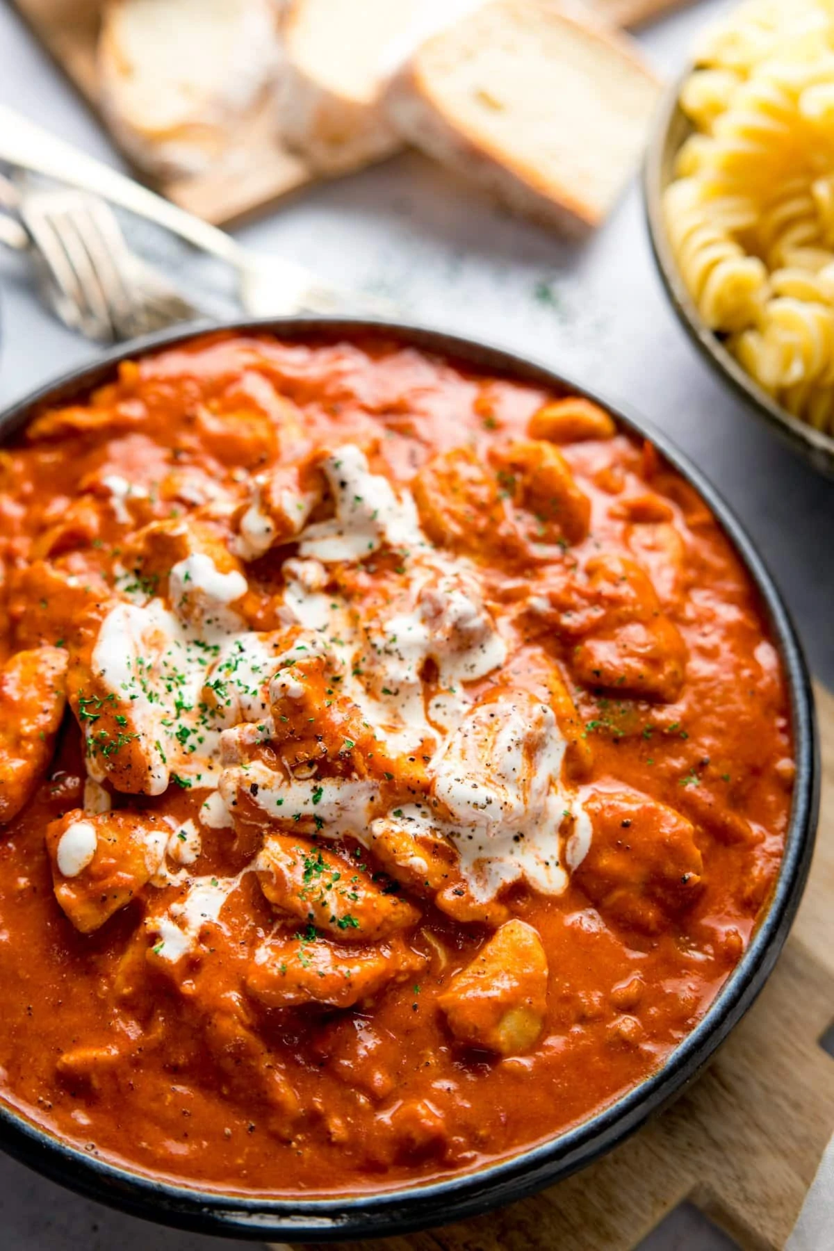 Dark bowl filled with chicken paprikash and topped with a swirl of soured cream. Bowl is on a light background and there are side dishes of bread and pasta just in shot.
