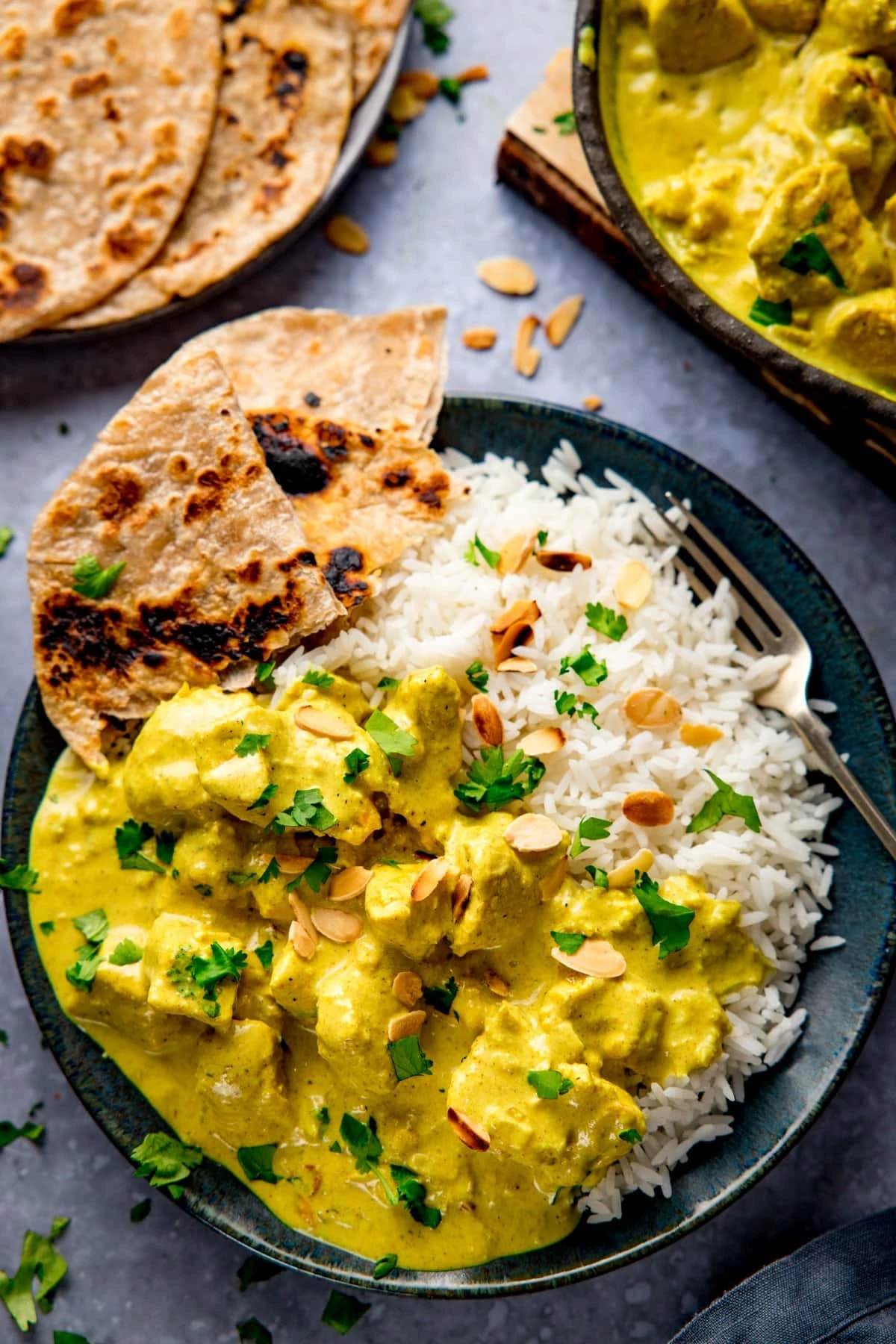 Dark plate with rice, chapati and chicken pasanda - topped with almonds and coriander. All on light blue background with further dishes just in shot at the top of the frame.