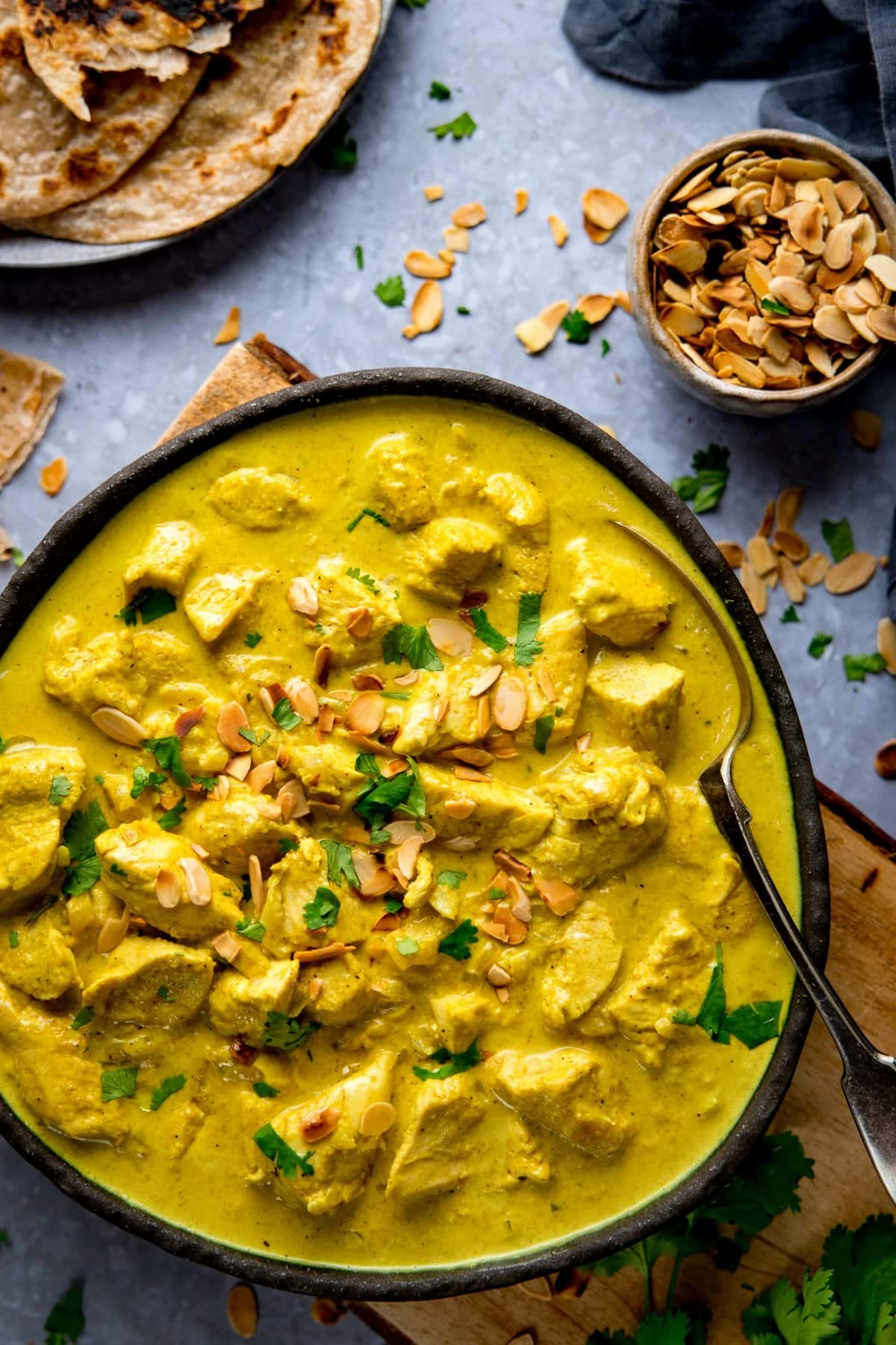 Dark bowl filled with Chicken pasanda. Bowl is sitting on a wooden chopping board on a light blue background. Further ingredients scattered around.