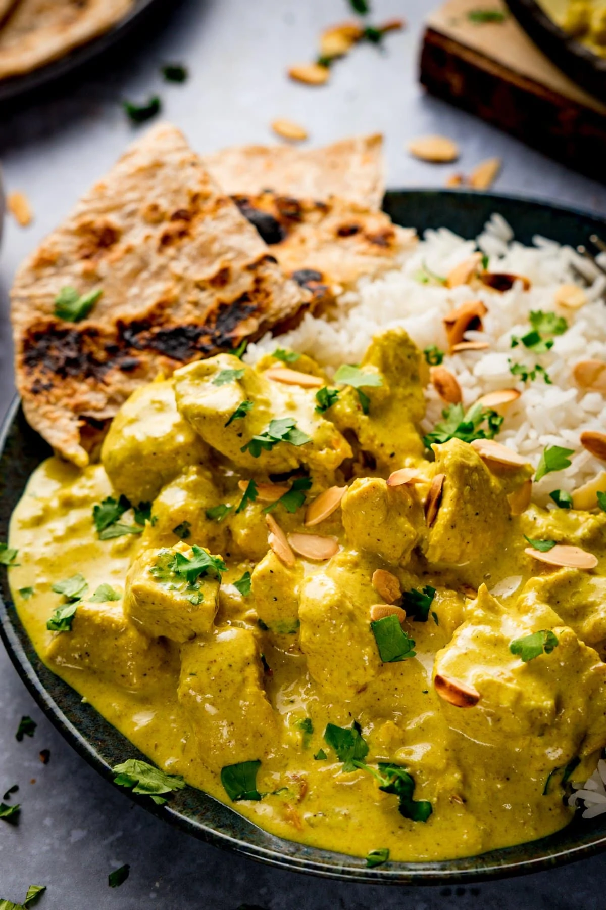 Dark plate with Chicken pasanda, rice and chapati on. Pasanda has coriander and toasted almonds sprinkled on top.