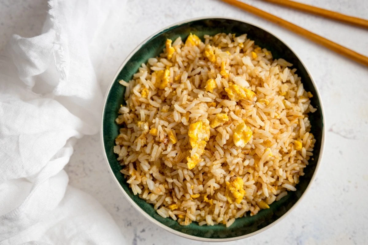 Wide image of egg fried rice in a dark bowl on a white table. White napkin and chopsticks on the table.