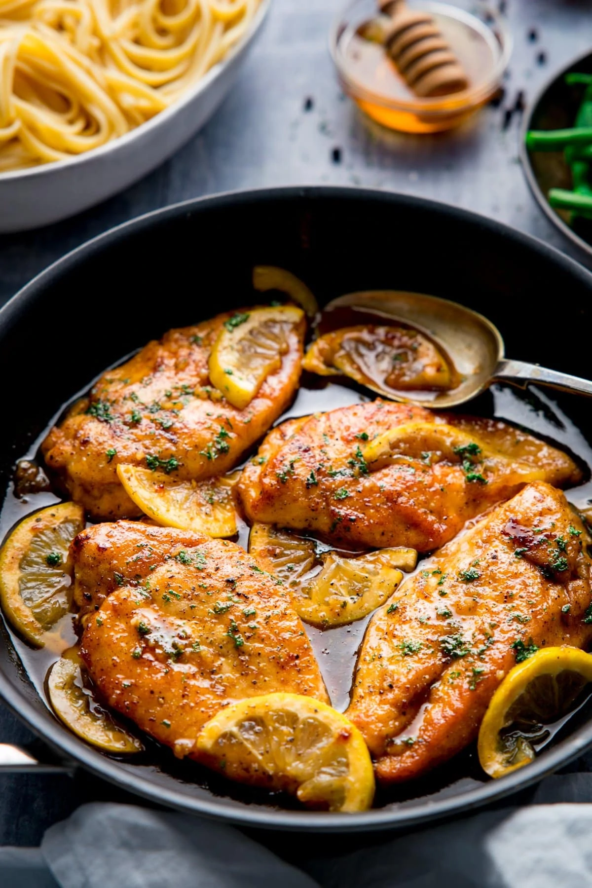 Black pan filled with honey lemon chicken on a blue background. Bowl of pasta and dish of honey just in shot.