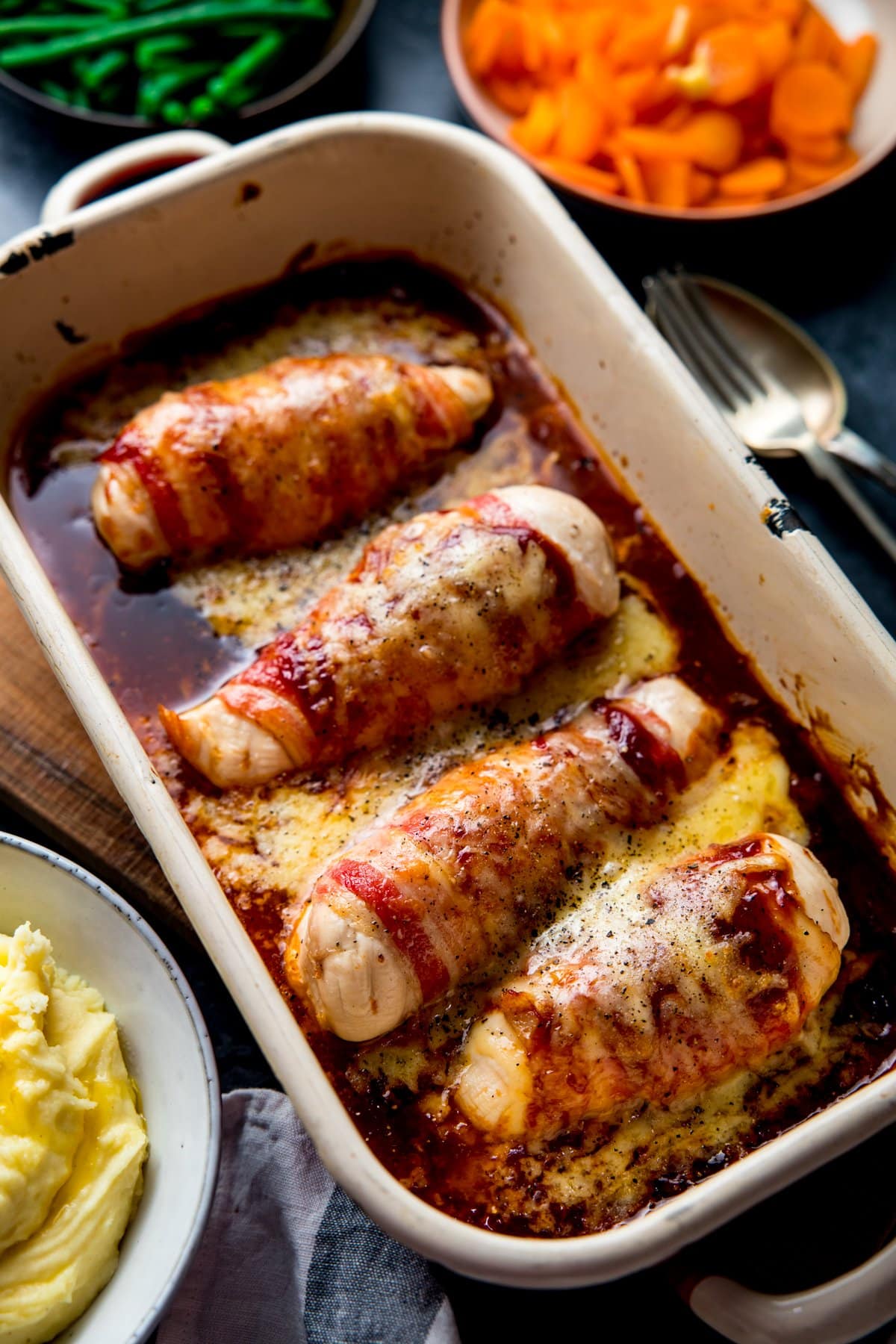 Hunters chicken (chicken wrapped in bacon with bbq sauce and cheddar) in a baking dish. Side dishes of carrots, green beans and mashed potatoes just in shot.