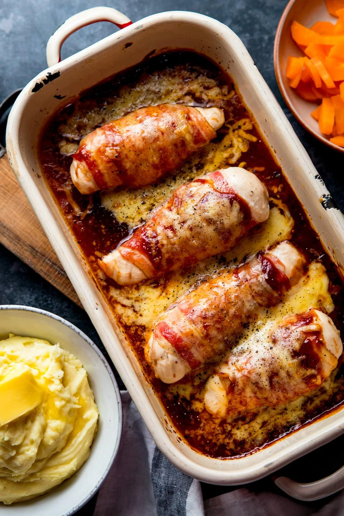 Hunters chicken (chicken with bacon, bbq sauce and cheese) in a baking tin on a dark surface. Side dishes of carrots and mashed potatoes are nearby.