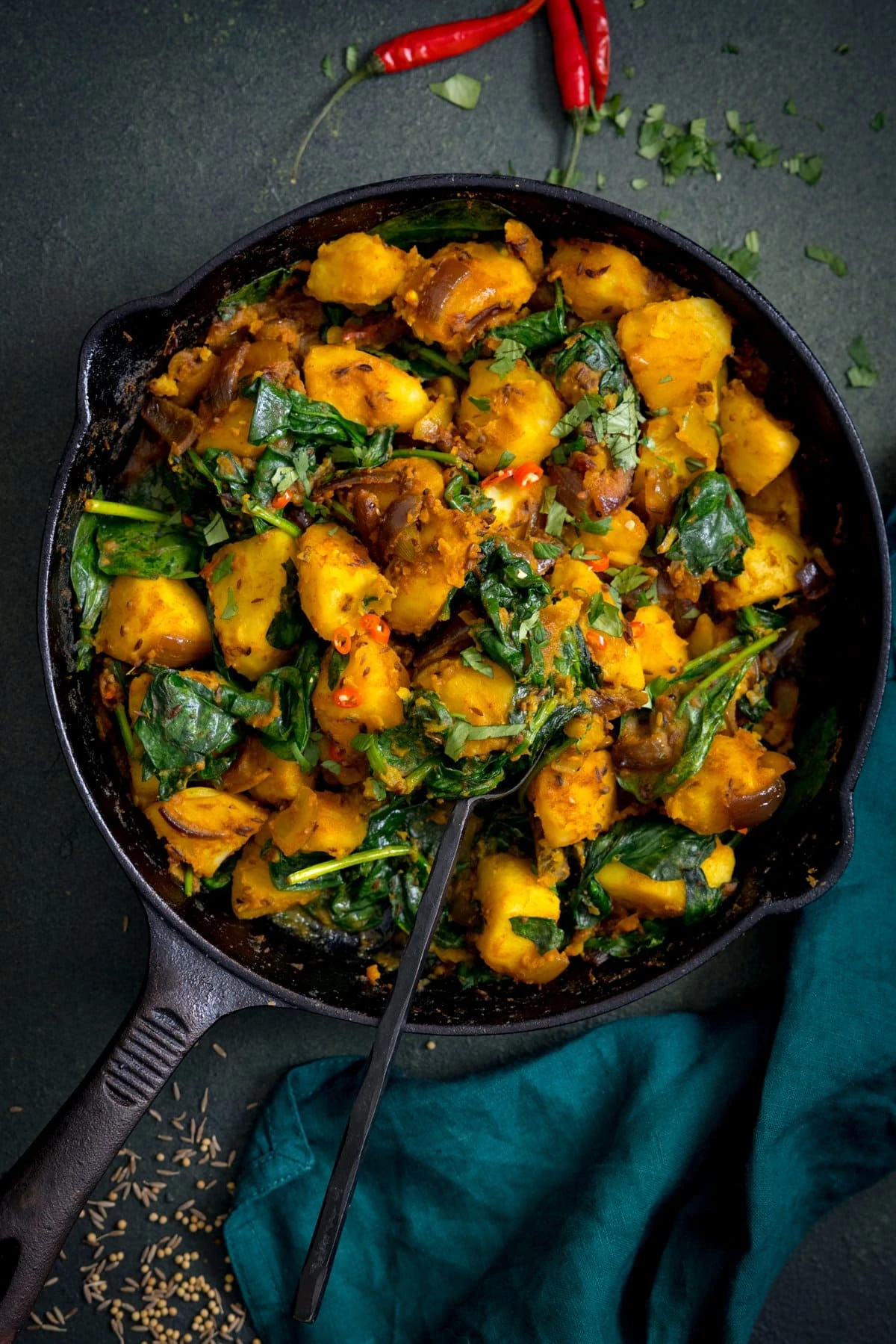 A spoonful of saag aloo being taken from a pan. The pan is on a dark green background.