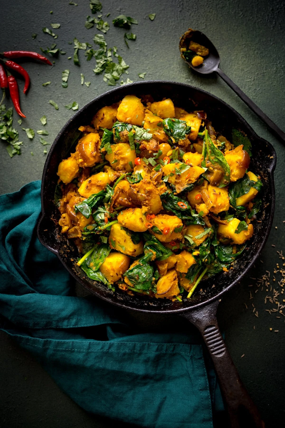 saag aloo in a cast iron pan on a dark green background/ Green napkin, a black spoon and garnishes surround the pan.