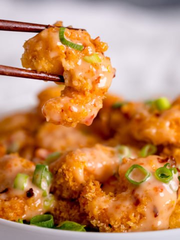 Crispy shrimp being lifted out of a bowl with chopsticks. Shrimp is covered with bang bang sauce.