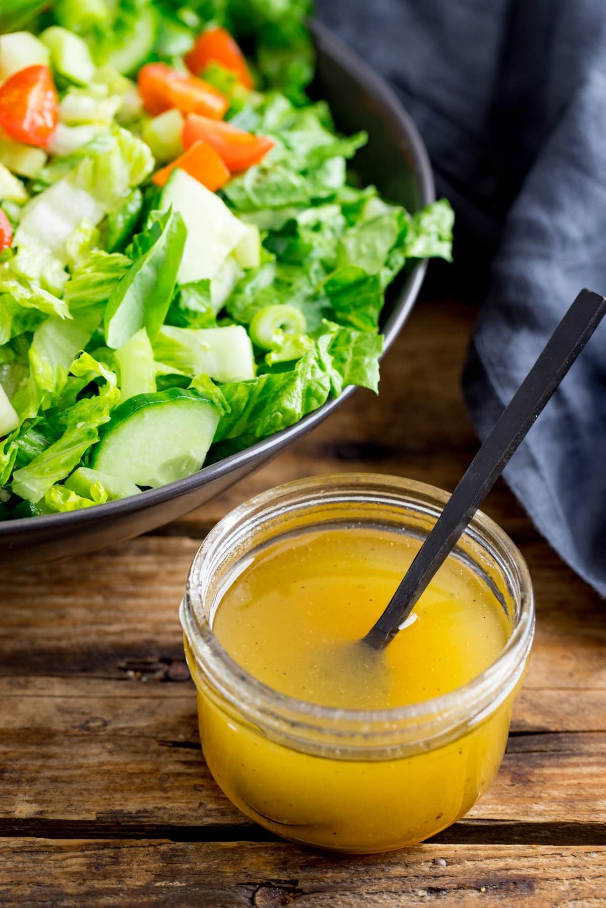 Jar of honey mustard salad dressing with a black spoon sticking out on a wooden table. Bowl of salad in the background.
