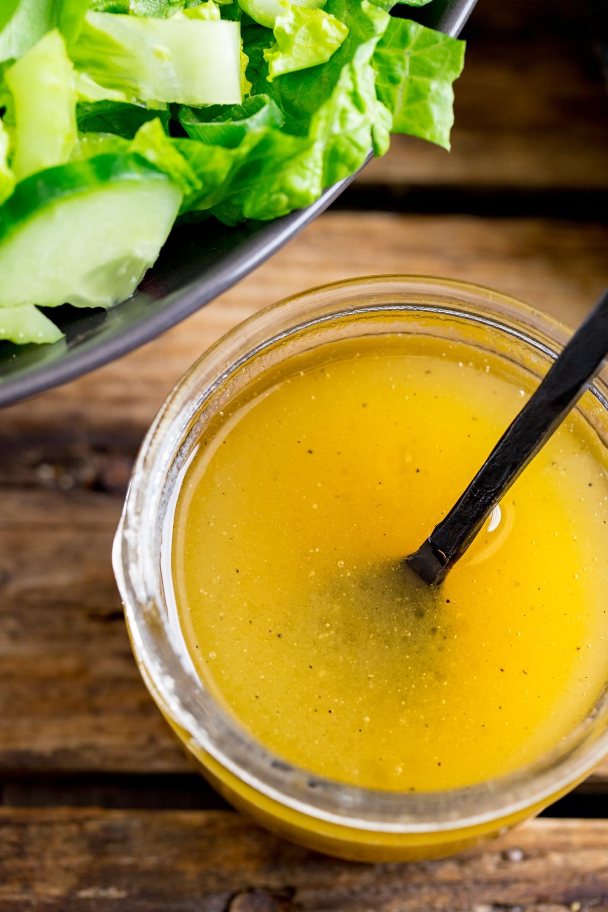 Overhead shot of a jar of honey mustard salad dressing on a wooden table. Bowl of salad partially in shot.