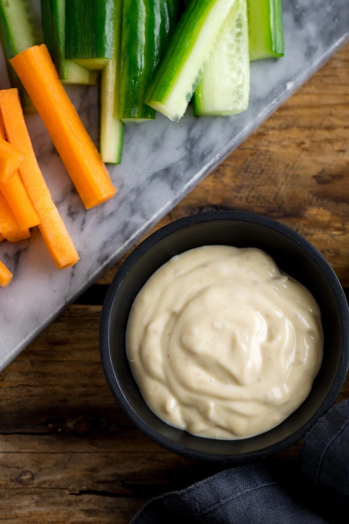 Overhead image of a small black bowl filled with salad cream next to carrot and cucumber pieces.