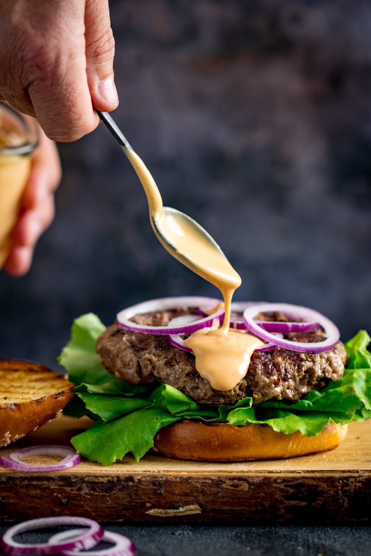 Burger sauce being drizzled from a spoon onto an open burger.