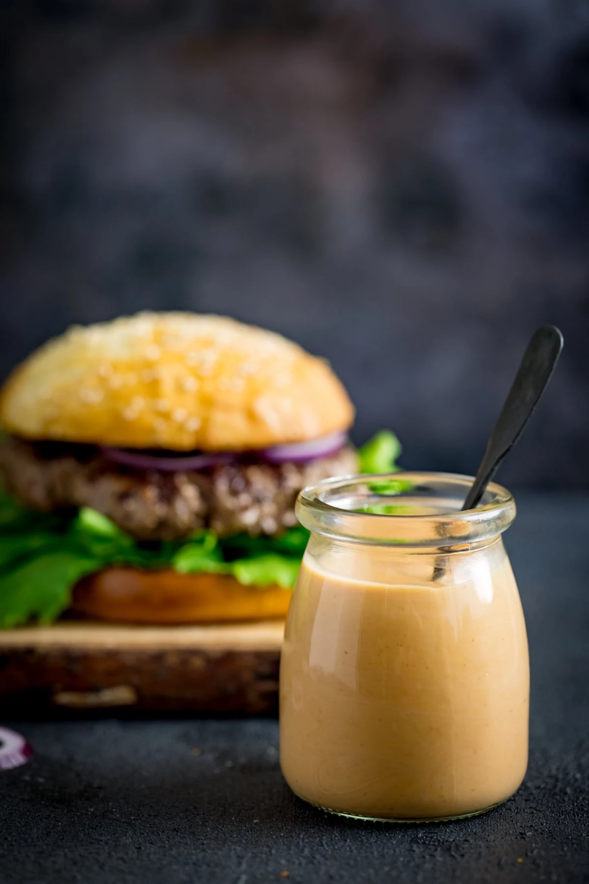 Burger sauce in a glass jar against a dark background. There is a burger in the background.