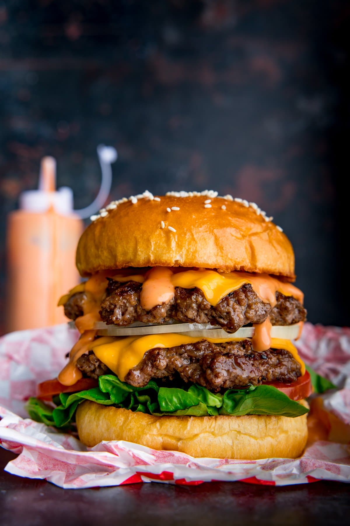 Double cheeseburger in an open wrapper on a dark background. Burger sauce in a bottle in the background.