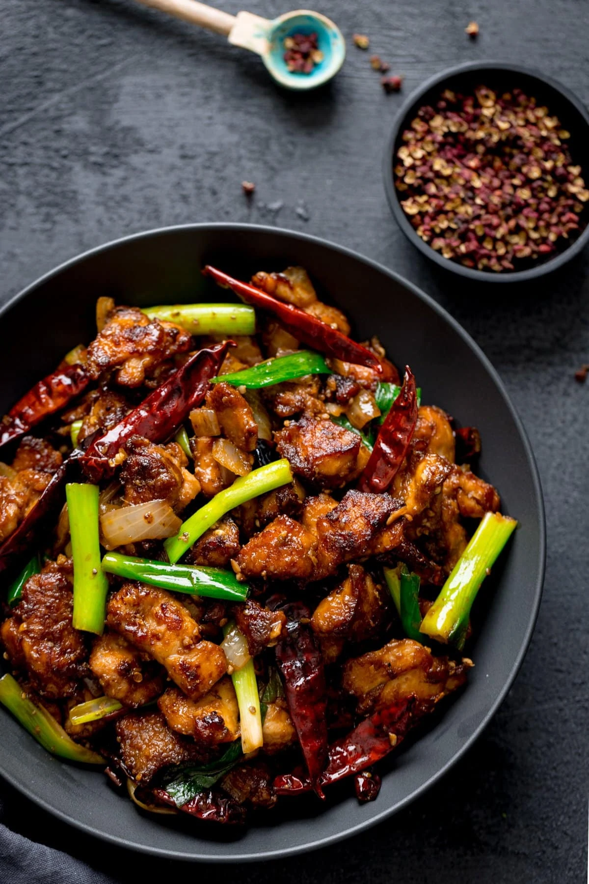 Szechuan chicken stir-fry in a black bowl on a dark surface. Little spoon and bowl of szechuan peppers in shot