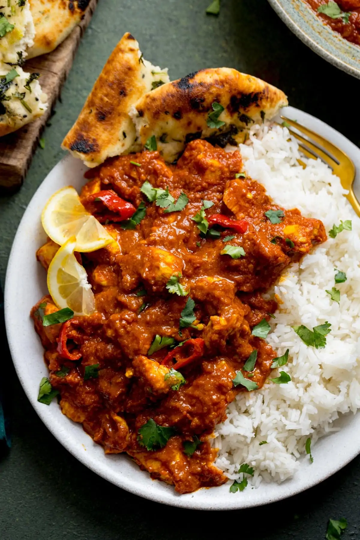 Chicken pathia with boiled rice and lemon slice son a white plate with naan bread. Plate is on a dark background,