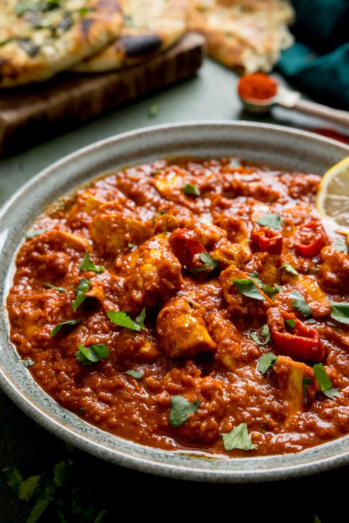 Chicken pathia in a green bowl on a dark background. Naan bread on a board just in shot at the top of the frame.