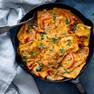 Overhead shot of ravioli in creamy tomato sauce in a black pan on a blue surface. There is a spoon in the pan and a light blue napkin next to the pan.