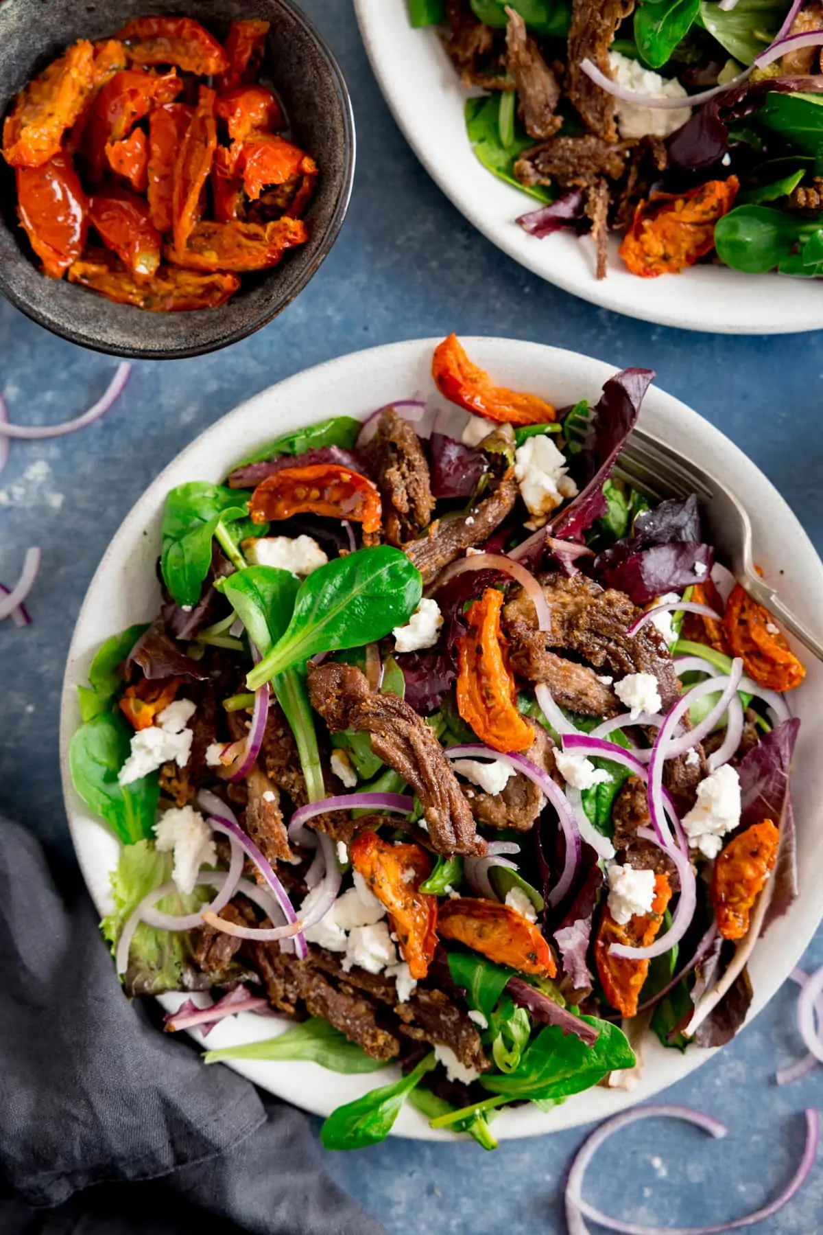 White plate filled with lamb, tomato, feta and red onion salad on a blue background. Further plate of salad and dish of tomatoes also in shot.