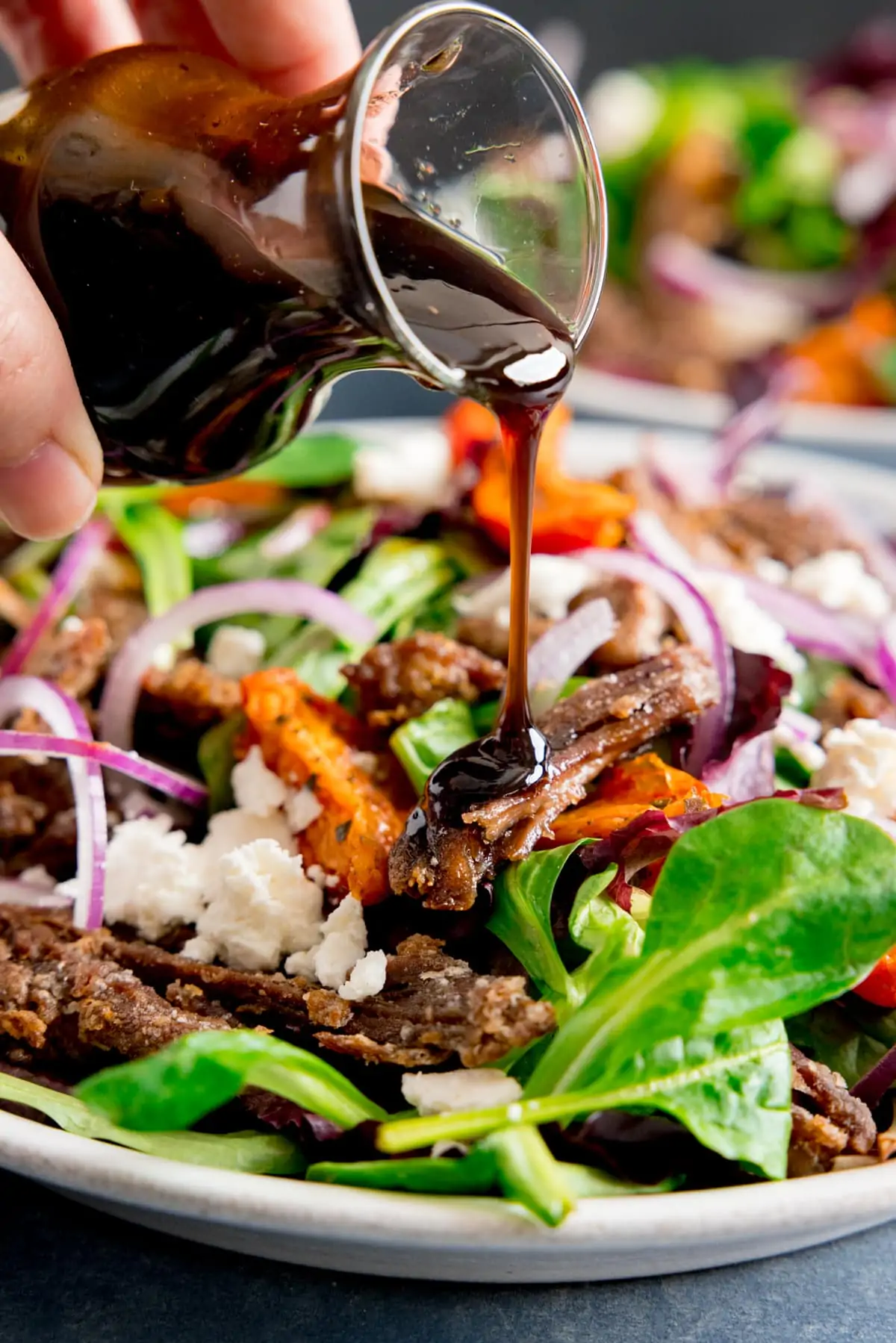 Close up of balsamic dressing being poured from a glass jar onto a crispy lamb, tomato and feta salad.