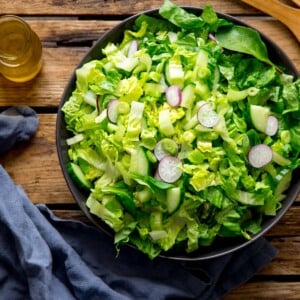 Large bowl of green salad on a wooden surface. Blue napkin and jar of salad dressing is next to the bowl.