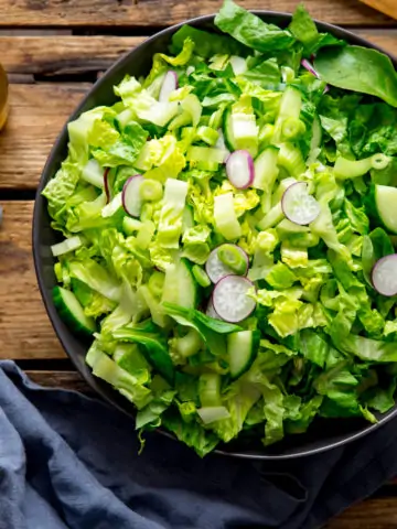 Large bowl of green salad on a wooden surface. Blue napkin and jar of salad dressing is next to the bowl.