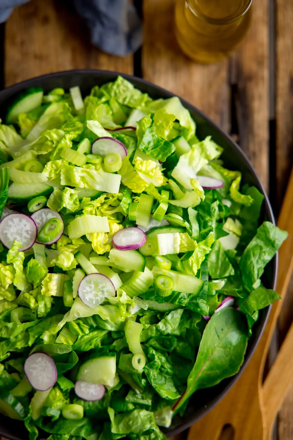 Overhead image of a bowl of simple green salad on a wooden table. Salad servers, salad dressing and a napkin are also in shot.