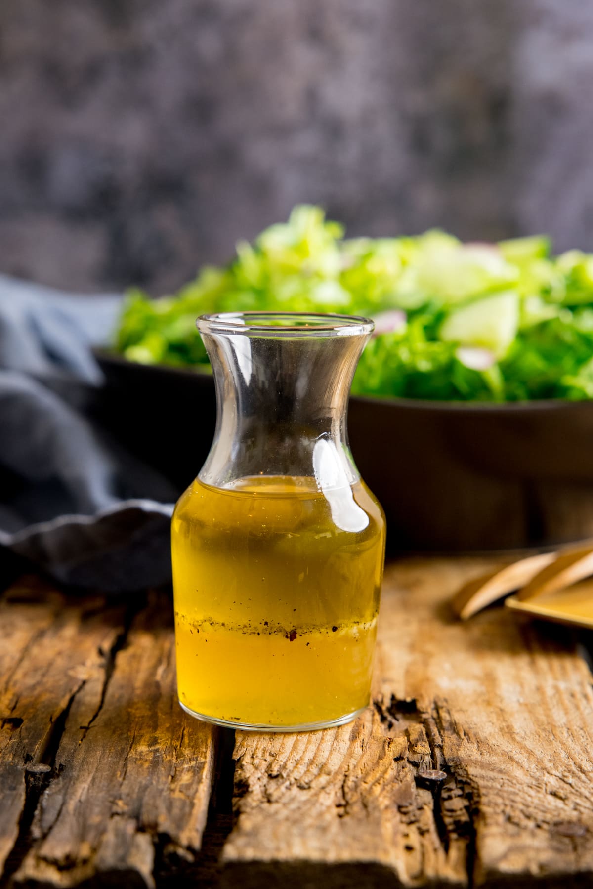Small glass jar filled with vinaigrette dressing on a wooden table. In the background is a bowl of salad and a blue napkin.
