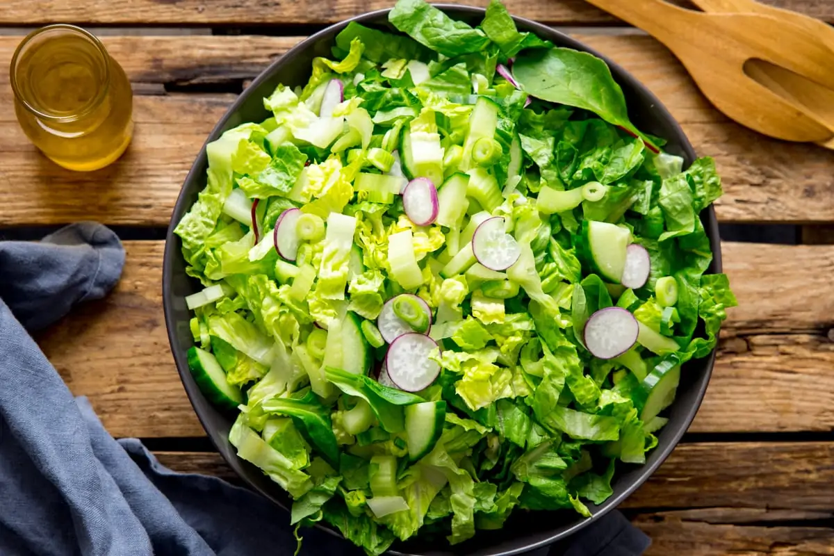 Wide image of a dark bowl filled with green salad on a wooden table. A blue napkin, jar of salad dressing and some wooden salad servers are next to the bowl.