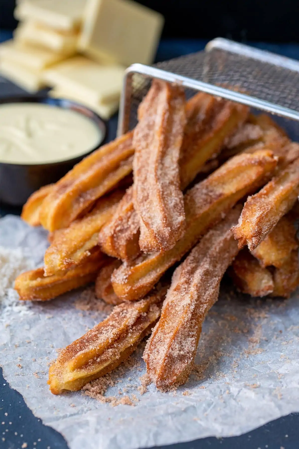 Picture of baked churros dusted with cinnamon sugar on their side in a wire basket with white chocolate in the background out of focus.