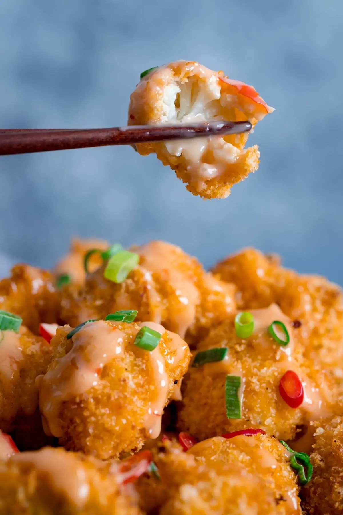 Piece of bang bang cauliflower with a bite out of it being lifted by chopsticks from a plate of bang bang cauliflower