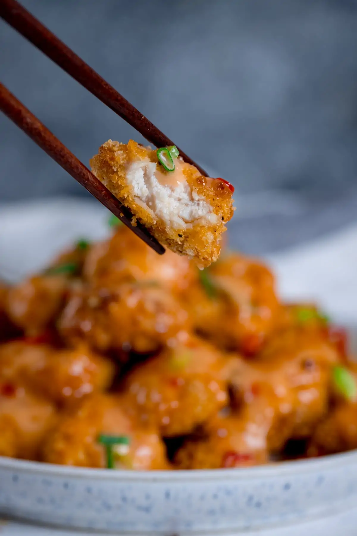 Piece of bang bang chicken with a bite out being held with a pair of chopsticks. Bowl of bang bang chicken in the background.