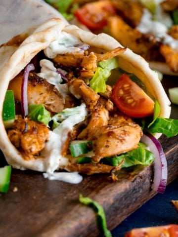 side shot of chicken gyros rolled up in a flatbread with lettuce, tomato, red onion and tzatziki. The gyros is on a dark wooden board with further ingredients scattered around.