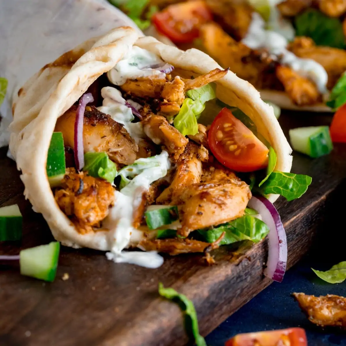 side shot of chicken gyros rolled up in a flatbread with lettuce, tomato, red onion and tzatziki. The gyros is on a dark wooden board with further ingredients scattered around.