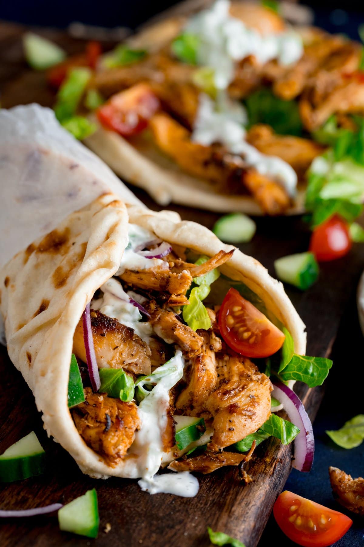 Chicken gyros in a rolled up flatbread with tzatziki and salad on a wooden board. Further ingredients and an unrolled gyros flatbread (with toppings) also in shot.