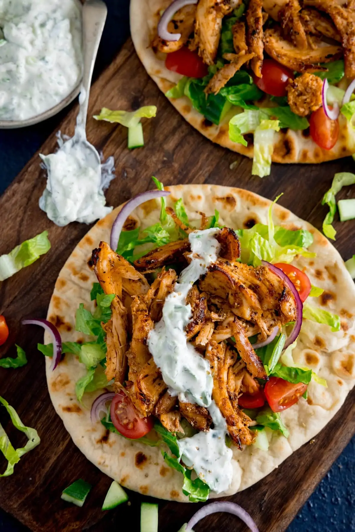 Chicken gyros with salad and tzatziki on an unrolled flatbread, on a wooden board. A further gyros flatbread and a bowl and spoon with tzatziki on is also in shot.
