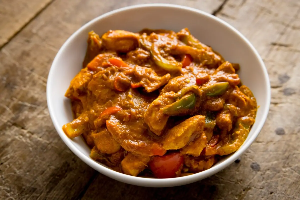 White bowl filled with chicken jalfrezi that has been made using curry base gravy. Bowl is on a wooden table.