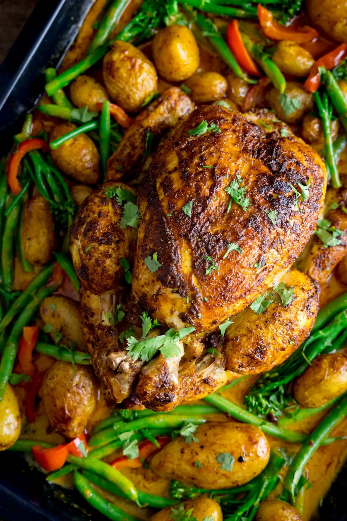 Overhead of a whole roast chicken in a baking tray with potatoes, peppers, green beans, broccoli and curry sauce