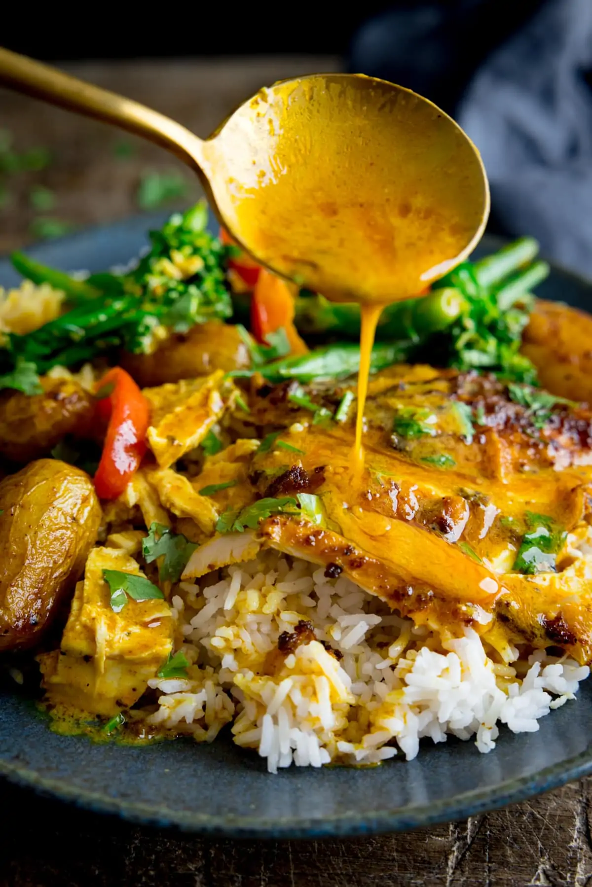 Curry sauce being spooned onto slices of curry roast chicken which is on a plate with rice and vegetables.