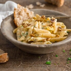 Square image of gorgonzola pasta in a light grey bowl on a wooden table. There is a piece of torn bread in the bowl with the pasta.