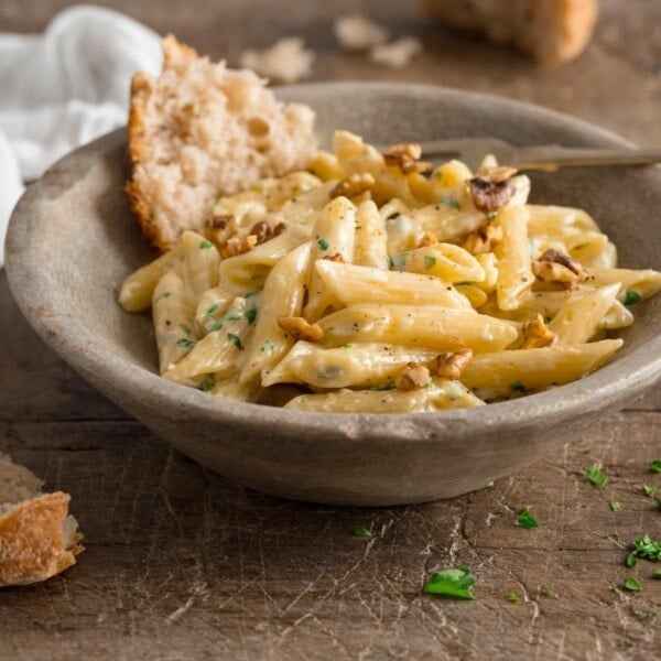 Square image of gorgonzola pasta in a light grey bowl on a wooden table. There is a piece of torn bread in the bowl with the pasta.