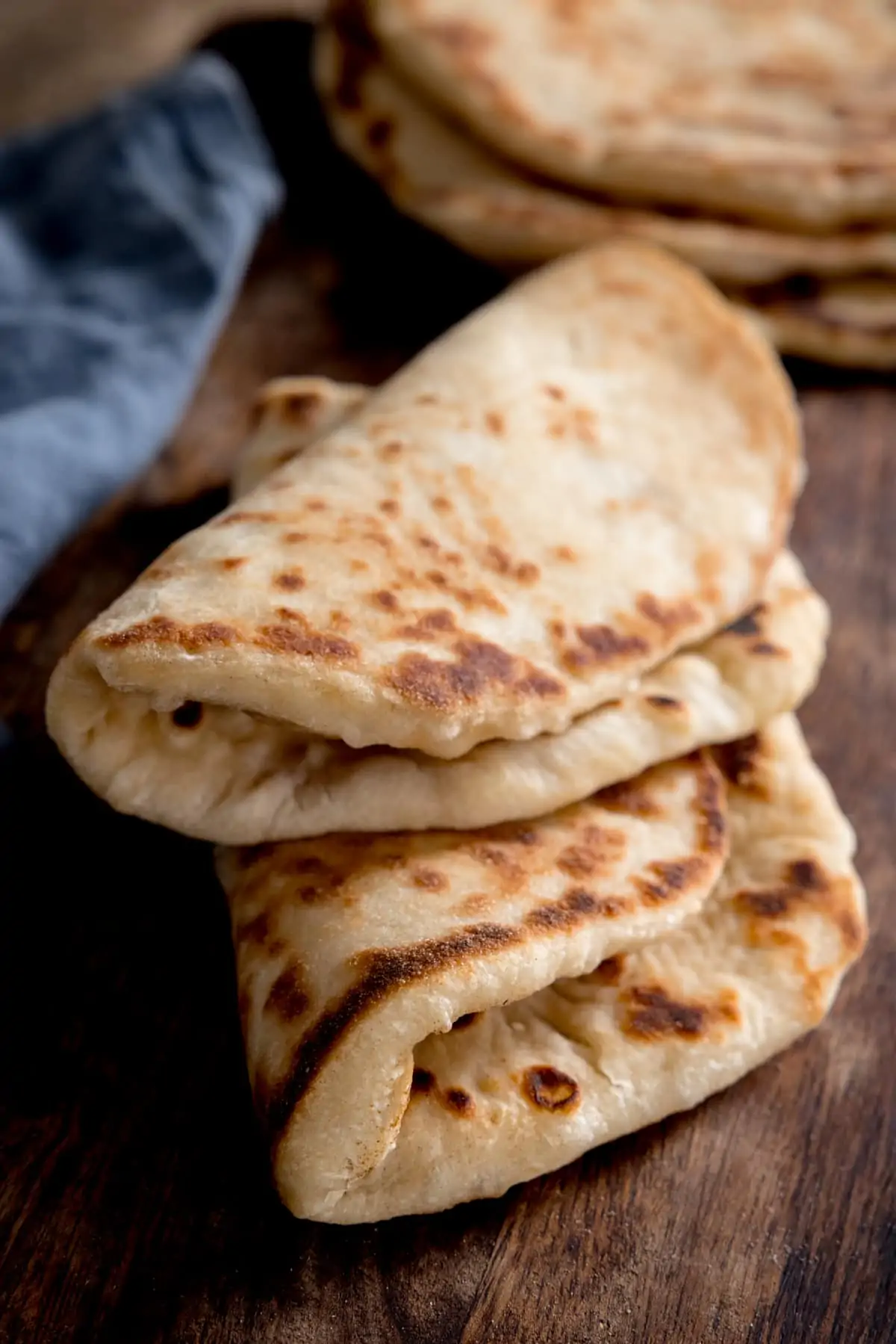 Two stacked folded flatbreads on a wooden board next to a blue napkin. Further flatbreads are in the background.