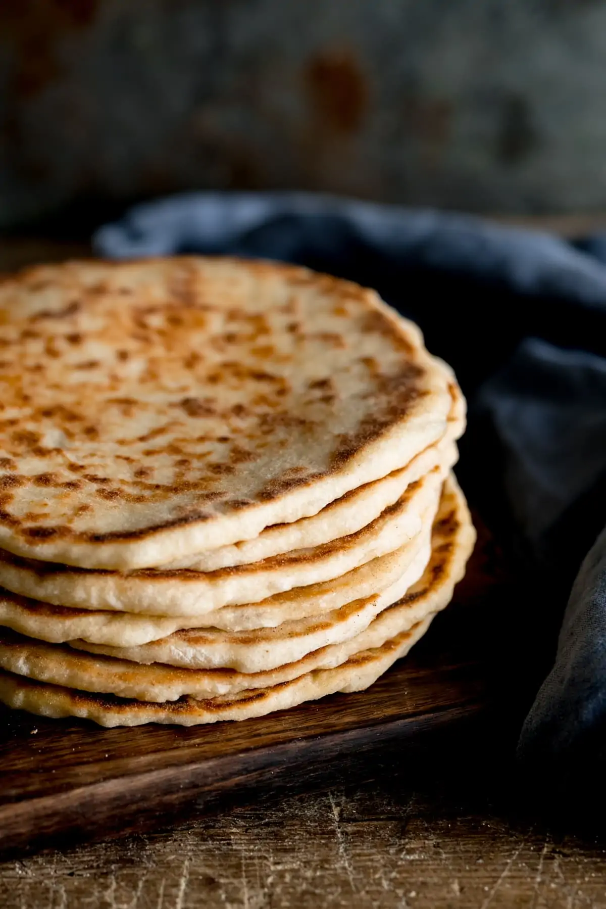 A stack of flatbreads on a wooden board against a dark background, next to a blue napkin.