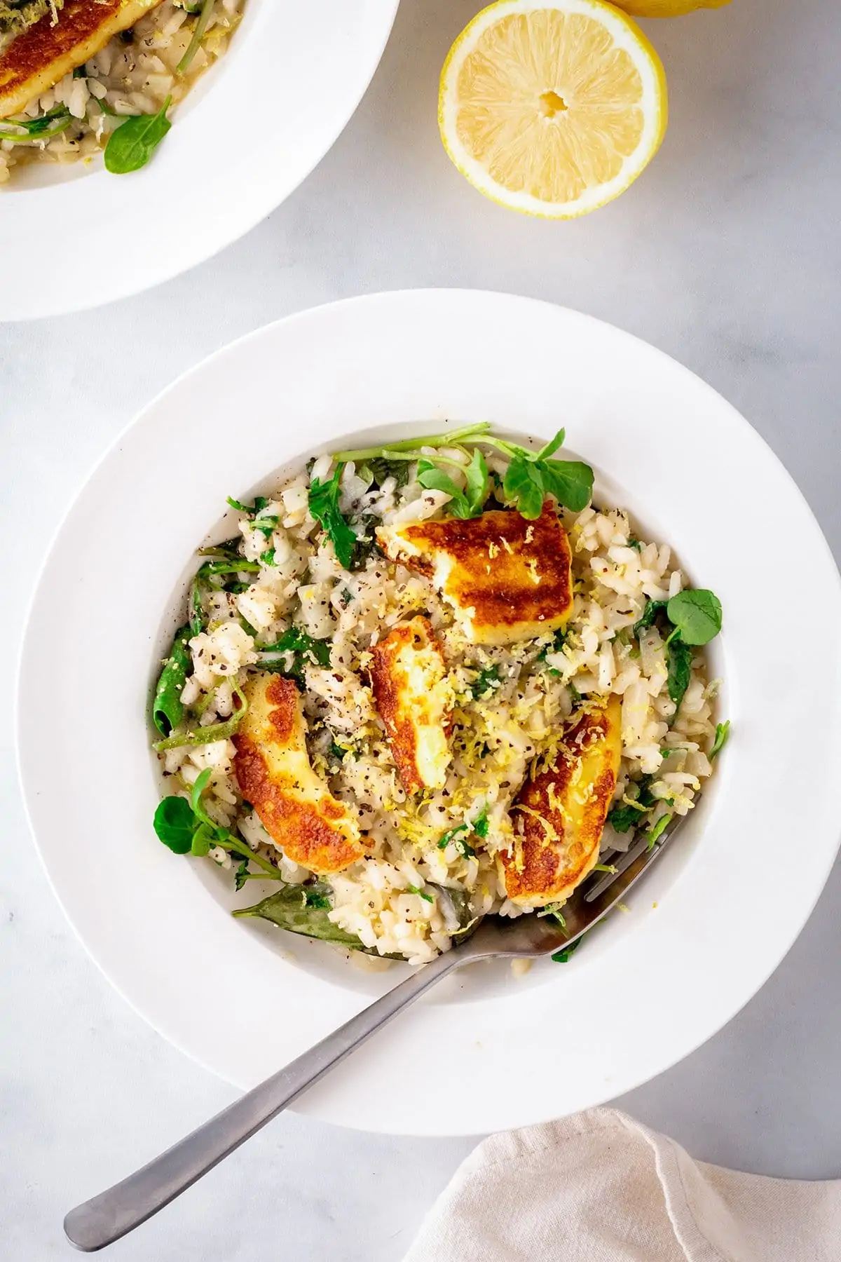 A white bowl with cooked lemon risotto topped with pieces of fried halloumi and a slice of lemon in the background.