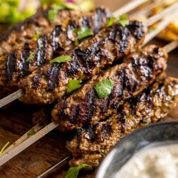 Close up pictures of lamb koftas on metal skewers sat on a wooden board with a bowl of dipping sauce in the foreground out of focus.