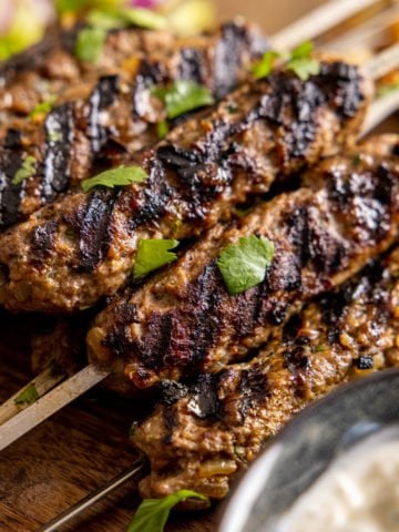 Close up pictures of lamb koftas on metal skewers sat on a wooden board with a bowl of dipping sauce in the foreground out of focus.