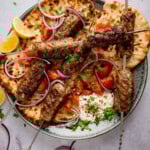 Grilled lamb kofta skewers on flatbread with sliced red onions, cherry tomatoes, lemon wedges, fresh herbs, and a dollop of whipped feta on a plate.