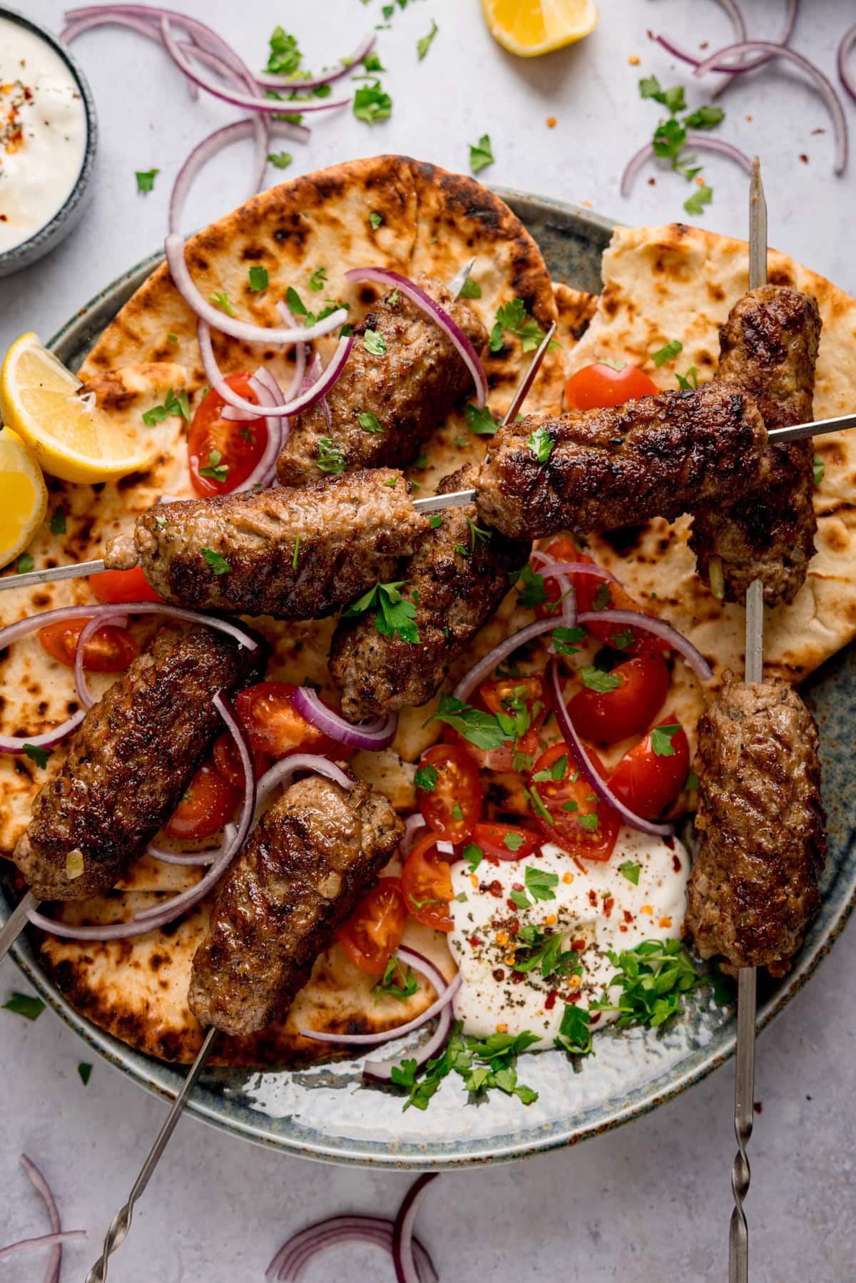 Overhead tall image of grilled lamb kofta skewers on flatbreads, on a plate, with sliced tomatoes, red onions, lemon wedges, fresh herbs, and a side of whipped feta.
